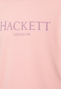 Jasnoróżowa bluza z bawełny, z nazwą marki "HACKETT" w fioletowych literach oraz napisem "LONDON" poniżej, mniejszą czcionką.