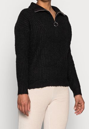 Persoon draagt een zwarte ribgebreide trui met een halve rits en een ringtrekker, gecombineerd met lichtbeige ribfluwelen broek.