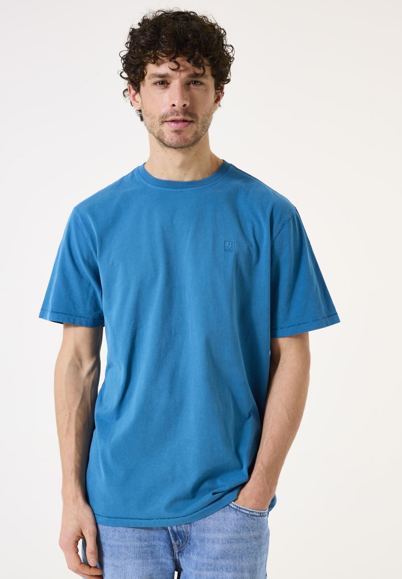 Türkises Baumwoll-T-Shirt mit Rundhalsausschnitt, kurzen Ärmeln und einem kleinen Logo-Patch auf der Brust. Getragen mit hellblauen Jeans.