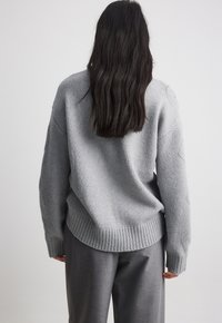 Pull en maille gris clair avec ourlet côtelé, épaules tombantes et manches longues ; porté sur un pantalon gris. Texture douce avec une coupe décontractée.