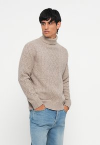Bežinis megztas turtleneckas su tekstūrine deimantine raštu, su raukiniais ant rankovių ir apykaklės, derinamas su šviesiai mėlynomis džinsais.