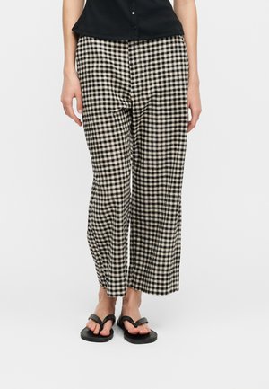 Femeie purtând pantaloni evazați cu imprimeu în carouri alb-negru, top negru fără mâneci și sandale negre tip papuc, stând pe un fundal alb.