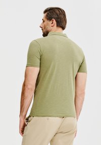 Koszulka polo z krótkim rękawem w oliwkowym kolorze, miękka w dotyku, klasyczny kołnierzyk i dopasowany krój. Noszona z jasnobeżowymi spodniami o prostym wzorze.