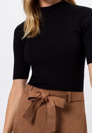 Vrouw die een zwart geribd T-shirt met korte mouwen draagt, ingestopt in een hoogtaillesuède broek in bruin met een strikceintuur.