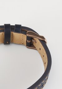 Bracelet de montre en cuir noir avec un dessous beige et une boucle en bronze sur fond blanc, enroulé en cercle.