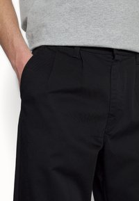 Personne portant un pantalon noir, les mains dans les poches, avec une chemise à manches courtes gris clair visible à la taille.