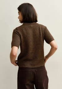 Femme avec des cheveux courts et foncés portant un haut en tricot marron à manches courtes et un pantalon marron foncé, se tenant avec les mains sur les hanches contre un mur uni.