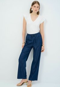 Pantalones de pierna ancha azules con una cinturilla elástica, combinados con una camiseta blanca ajustada de cuello en V. El modelo lleva sandalias amarillas con tiras.