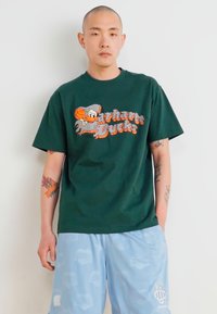 T-shirt en coton vert avec un graphique coloré "Bucks" et un ballon de basket, associé à un short bleu clair présentant un motif de logo répétitif.