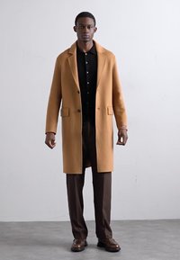Lichtbruin wollen jasje met een notchrevers, enkelvoudige knopen, gecombineerd met donkere broeken en een zwart shirt.