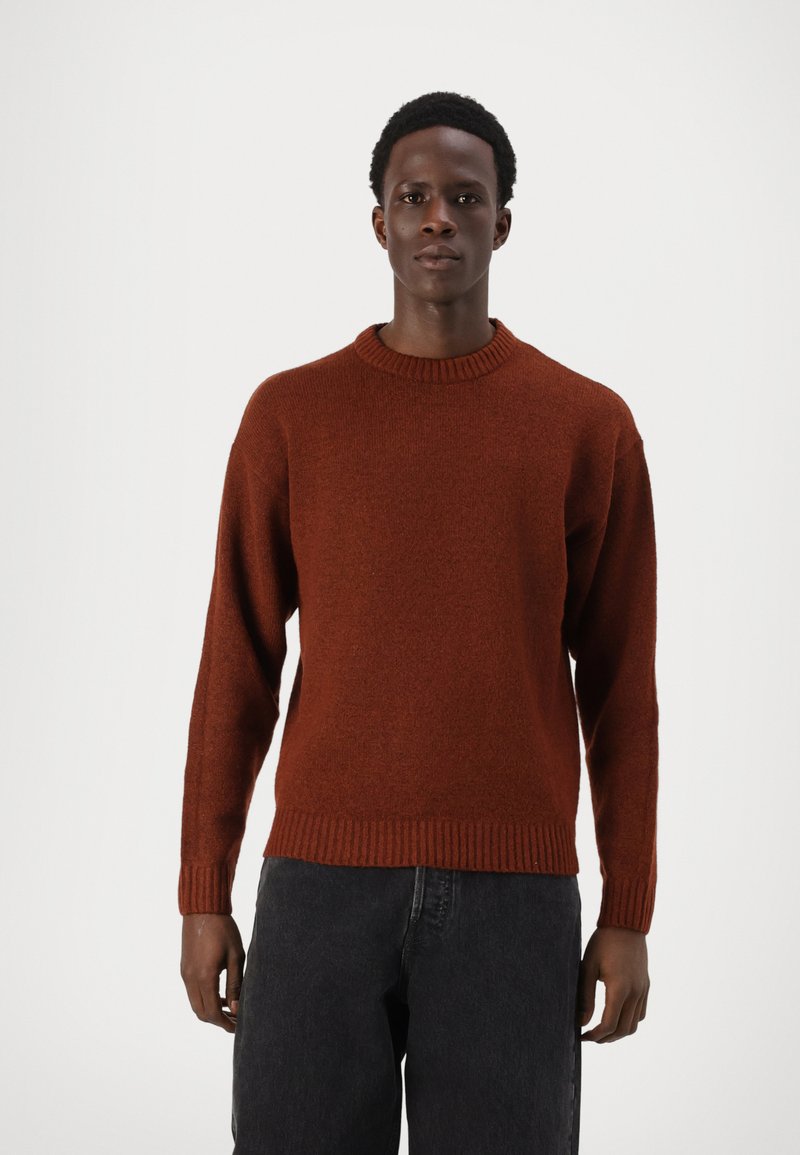 Pull en tricot couleur rouille avec un col rond, des poignets et un ourlet côtelés, présentant une texture douce. Porté avec un pantalon sombre.