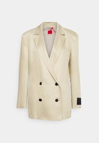 Blazer beige de doble botonadura con solapas prominentes, dos botones negros y una etiqueta de tela en la manga izquierda. Textura suave.