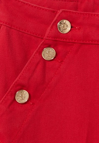 Tela de mezclilla roja con una textura suave, que presenta botones de metal dorado y costuras en contraste a lo largo de la cremallera y la cinturilla.