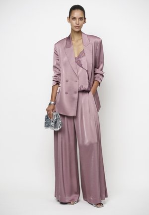 Vrouw draagt een mauvekleurig satijnen pak met oversized blazer, broek met wijde pijpen, top met kanten rand, houdt een zilveren pailletten clutch vast.