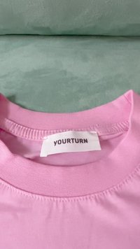 Tricou roz din bumbac cu guler rotund tricotat, având o etichetă albă pe care scrie "YOURTURN". Textură netedă și detalii cusute.