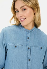 Lichtblauwe denim overhemd met een hoge kraag, vooraan knoopsluiting en twee borstzakken met bruine knopen. Soepele textuur en casual ontwerp.