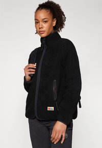 Giacca in pile nera con colletto alto, tasche con zip e patch del logo. Presenta una superficie morbida e textured e una vestibilità leggermente over-size.