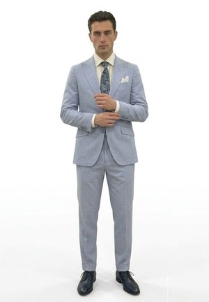 Hombre ajustando los puños, vestido con traje azul claro, camisa blanca, corbata azul con patrón, pañuelo blanco en el bolsillo y zapatos de vestir negros, sobre fondo blanco.