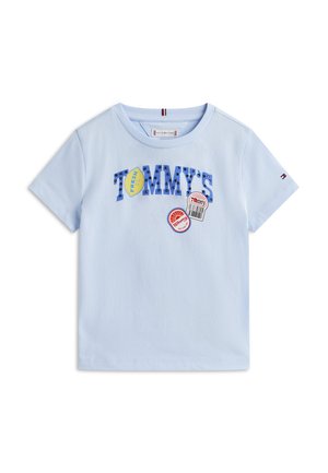 Hellblaues T-Shirt mit kurzen Ärmeln, auf dem "TOMMY'S" in blauen Nieten steht, sowie bunten Aufnähern, darunter ein gelbes Hexagon mit der Aufschrift "FRESH" und ein Barcode-Design.