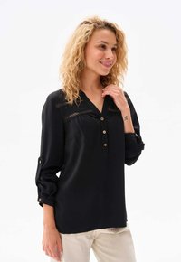 Schwarze Bluse mit langen Ärmeln und Knopfleiste, die dekorative Spitzendetails über der Brust sowie aufrollbare Ärmel mit Knopf-Detail besitzt.