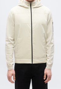 Homme portant une veste à capuche beige zippée avec poches avant et un pantalon noir sur fond uni.