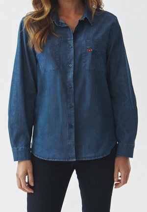 Frau trägt ein blaues Jeanshemd mit Knöpfen und Brusttaschen, eine Tasche ist mit kleinen roten Kirschen bestickt.