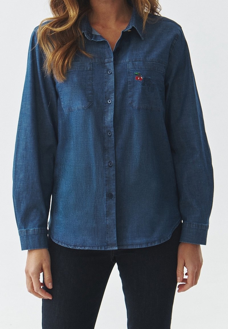 Femme portant une chemise en jean bleue avec des poches poitrine, une poche brodée de petites cerises rouges.
