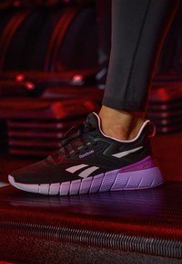 Pie con zapatilla Reebok negra y blanca pisando una plataforma texturizada negra en una iluminación roja tenue, el fondo incluye pesos apilados.