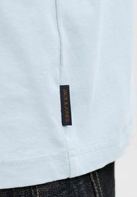 Ανοιχτό μπλε βαμβακερό μπλουζάκι με ίσια εσοχή, διαθέτοντας μια επώνυμη ναυτική ετικέτα με την ένδειξη "JACK & JONES" σε πορτοκαλί στη ραφή της πλευράς.