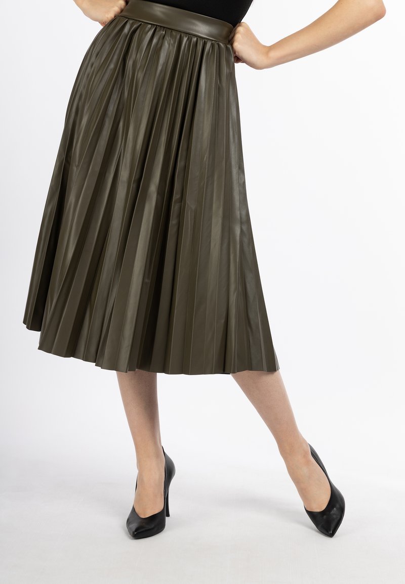 Olijfgroene, geplooide faux leren rok met een brede tailleband, gecombineerd met zwarte hoge hakken tegen een witte achtergrond.