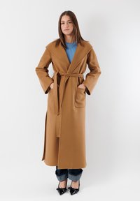 Cappotto lungo color cammello con ampi rever, vita con cintura e tasche frontali. Il modello indossa un maglione blu e jeans scuri con orli risvoltati e tacchi neri.