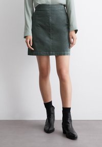 Falda mini de mezclilla verde con corte recto, que presenta un brillo sutil y dobladillo estándar. Combinada con botas negras de tobillo y calcetines.