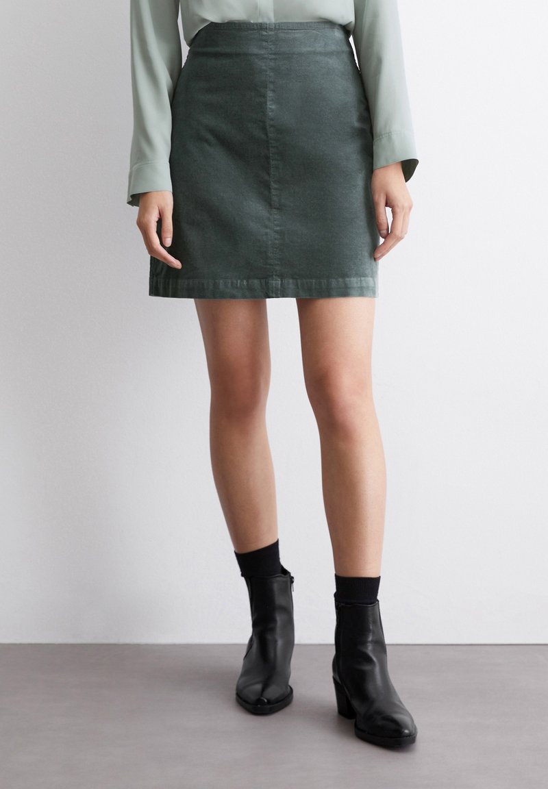 Falda mini de mezclilla verde con corte recto, que presenta un brillo sutil y dobladillo estándar. Combinada con botas negras de tobillo y calcetines.