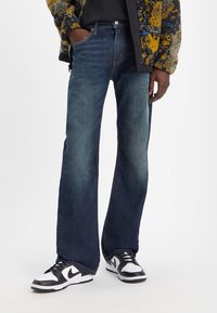 Mörkblå denimjeans med utsvängd form, med en framknapp och dragkedja, bärs med en svart topp och svart-vita sneakers.
