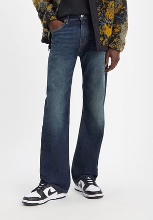 Jeans de mezclilla de color azul oscuro con forma acampanada, con un botón y cremallera en el frente, combinados con una camiseta negra y zapatillas blanco y negro.