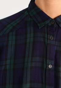 Chemise à carreaux pour hommes avec une combinaison de couleurs marine et verte, col pointu, fermeture à boutons et tissu texturé avec des lignes verticales et horizontales.