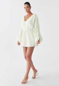 Model in een witte, geplooide mini-jurk met een diepe V-hals en volumineuze mouwen, gecombineerd met witte sandalen met banden, tegen een egaal achtergrond.