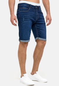 Bermudas de denim para hombre, de color azul oscuro con un acabado descolorido, dobladillos enrollados, diseño de cinco bolsillos, cierre con cremallera y acentos de costura clara.