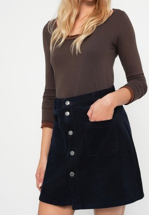 Femme portant un haut à manches longues marron foncé et une jupe en velours côtelé bleu marine boutonnée devant avec des poches, main dans une poche.