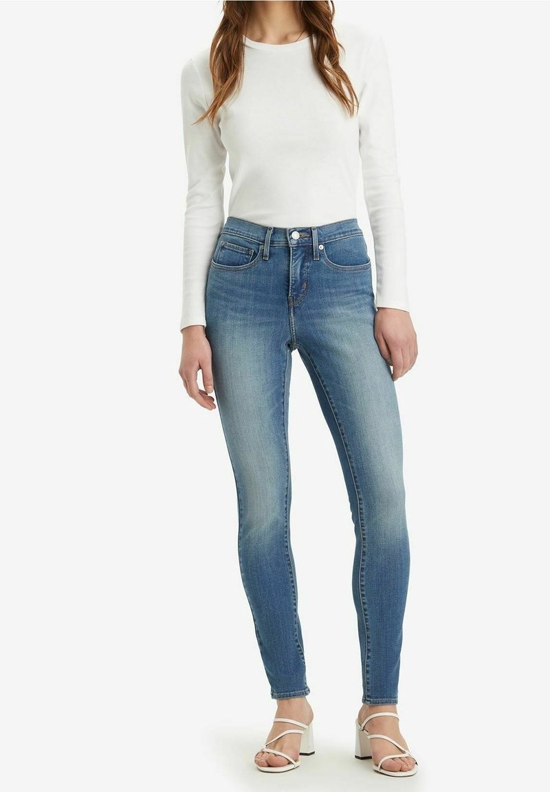 Femme portant un jean skinny taille haute bleu clair, un haut blanc à manches longues et des sandales blanches à talons avec des brides.