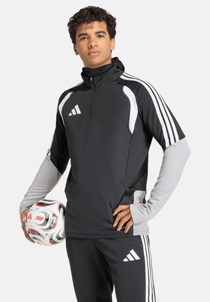 Mężczyzna ubrany w czarno-szaty strój sportowy Adidas, trzymający białą piłkę nożną Adidas Tiro w jednej ręce na jednolitym tle.