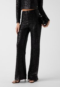 Schwarzes Pailletten-Zweiteiler-Outfit mit einem kurzen, langärmeligen Oberteil und ausgestellten Hosen. Das Ensemble hat eine glänzende Textur und eine figurbetonte Silhouette.