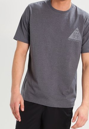 Mann trägt ein graues Under Armour T-Shirt mit kurzen Ärmeln und schwarze Shorts, steht mit entspannten Armen an den Seiten.