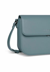 Taschen in Türkis aus Leder mit Klappenverschluss, verstellbarem Riemen und dezentem Branding auf der Vorderseite. Die Tasche hat eine glatte Textur und abgerundete Kanten.