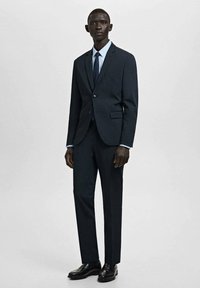 Donker marineblauw pak met een getailleerd colbert en rechte pijpen. Gecombineerd met een lichtblauw overhemd en een donkere stropdas, afgewerkt met zwarte schoenen.