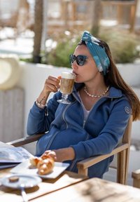 Felpa blu con zip realizzata in tessuto morbido, abbinata a una fascia per capelli a fantasia, occhiali da sole e gioielli di conchiglia; tiene un latte vicino a dei dolci.