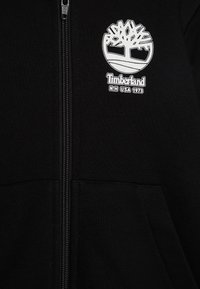 Čierna zapínacia mikina s kapucňou so svetlým logom Timberland a textom, vyrobená z mäkkej látky s hladkou textúrou a bočnými vreckami.