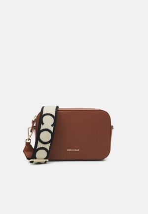 Brun texturerad läder crossbody-väska med svart och vit rem med stora kontrasterande bokstäver, guldhardware och varumärkeslogotyp på framsidan.
