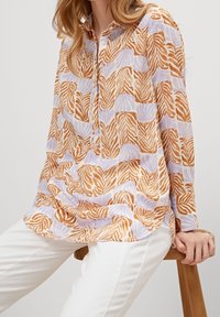 Blusa a maniche lunghe con colletto in un tessuto leggero, caratterizzata da un motivo di foglie arancioni, marroni e lavanda, abbinata a pantaloni bianchi.