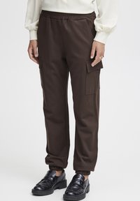 Bruine cargobroek met een elastische tailleband, zijzakken en geribbelde manchetten, gecombineerd met zwarte loafers met een decoratieve band.